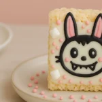 Cute Labubu-themed Rice Krispie treat decorated with white chocolate, pink sprinkles, and marshmallows on a ceramic plate.