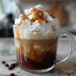Glass mug filled with iced cloud coffee topped with whipped cream and caramel drizzle, on a marble countertop