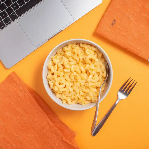 Bowl of creamy mac and cheese on an orange desk next to a laptop, fork, and napkins – easy student meal