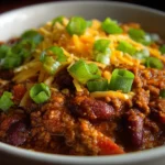 Hearty chili con carne topped with shredded cheddar and green onions in a white bowl