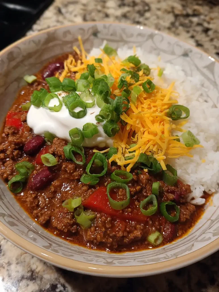 A bowl of chili con carne served with white rice, topped with shredded cheddar cheese, sour cream, and chopped green onions.