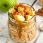 Caramel apple overnight oats in a glass jar topped with apple chunks and caramel drizzle on a white marble background with cinnamon sticks and a green apple – healthy budget-friendly student breakfast.