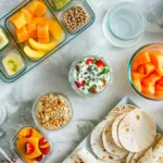 Overhead flat lay of easy dorm meal prep foods for college students, including overnight oats jars, tortilla wraps, fruit cups, and a microwave bowl near a mini fridge with snacks — bright and colorful no-cook meal prep setup.
