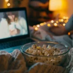Student in a cozy dorm room enjoying homemade microwave popcorn while watching a movie on a laptop.