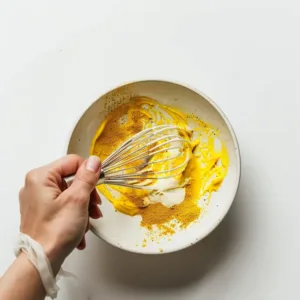 Mixing yogurt with turmeric and cumin in a small bowl on a white background.