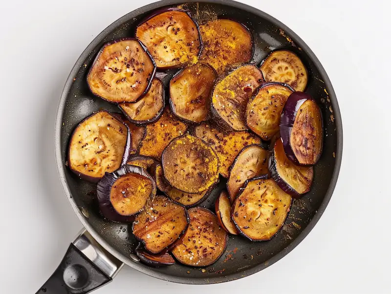 Sliced eggplants cooking in a skillet with turmeric and cumin on a white background.