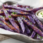 Crispy baked purple sweet potato fries on a parchment-lined tray with garlic aioli and chopped parsley, showing golden edges and vibrant purple color.