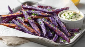 Crispy baked purple sweet potato fries on a parchment-lined tray with garlic aioli and chopped parsley, showing golden edges and vibrant purple color.