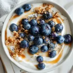 Top-down Greek Yogurt Blueberry Breakfast Bowl with honey, granola and chia on a white desk.