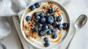 Top-down Greek Yogurt Blueberry Breakfast Bowl with honey, granola and chia on a white desk.