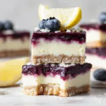 Keto blueberry lemon cheesecake bars stacked on a plate with almond flour crust, lemon cheesecake filling, blueberry layer, and coconut crumble topping.