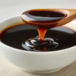 Dark honey garlic sauce in a white bowl with glossy soy-based texture, thick black garlic sauce dripping from a spoon