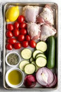 Easy Greek Sheet Pan Chicken Dinner (One-Pan Mediterranean Favorite) 5 Ingredients for Greek sheet pan chicken dinner including chicken thighs, cherry tomatoes, zucchini, red onion, garlic, lemon, olive oil, and oregano