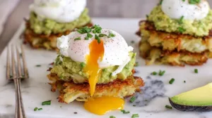 Potato Avocado Toast made with crispy shredded potato hash brown patties topped with mashed avocado and a perfectly poached egg with runny yolk on a white marble board.