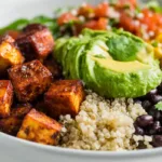A colorful vegan high protein burrito bowl filled with crispy tofu, quinoa, black beans, corn, avocado, tomatoes, and lime wedges on a white plate in bright natural light.