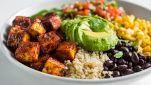 A colorful vegan high protein burrito bowl filled with crispy tofu, quinoa, black beans, corn, avocado, tomatoes, and lime wedges on a white plate in bright natural light.