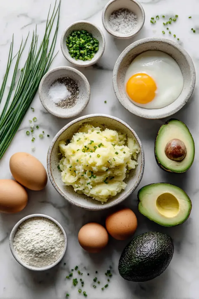 Ingredients for Potato Avocado Toast on a white marble surface including grated potatoes, avocado, eggs, cassava flour, arrowroot powder, chives, salt, and pepper.