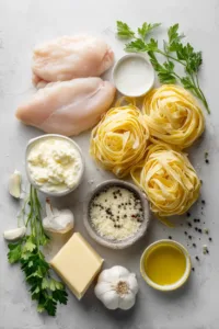Chicken Fettuccine Alfredo (Better Than a Restaurant) 5 Flat lay of ingredients for chicken fettuccine alfredo: raw chicken breasts, fettuccine pasta, garlic cloves, butter, parmesan cheese, heavy cream, and olive oil on a wooden table.