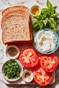 Flatlay of tomatoes, bread, cream cheese, herbs, olive oil, lemon, mayo, salt, and pepper on a marble board.