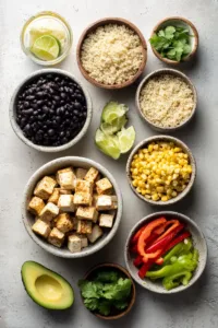 Top-down flatlay of vegan burrito bowl ingredients: tofu cubes, cooked quinoa, black beans, corn, avocado, bell pepper, lime, and cilantro arranged neatly on a light surface.