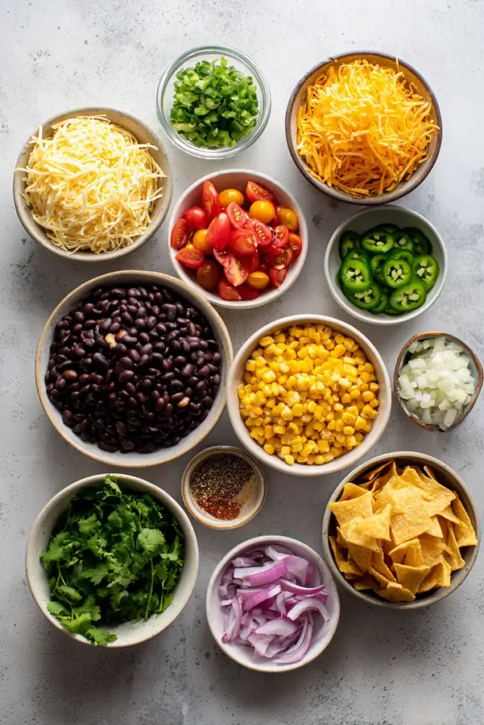 Cowboy Caviar Jalapeno Popper Cheese Log (Easy Party Appetizer) 3 Overhead flat lay of ingredients for cowboy caviar jalapeno popper cheese log including cream cheese, shredded cheddar, black beans, corn, jalapeños, red onion, tomatoes, cilantro, and tortilla chips in small bowls.