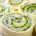 Cucumber and dill pinwheels sliced into rounds with visible spiral layers, cream cheese filling and thin cucumber strips inside soft flour tortillas on a white platter