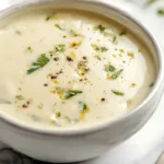 Creamy garlic parmesan sauce in a white bowl topped with parsley and black pepper on a light marble countertop.
