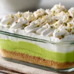 Layered pistachio dessert in a glass baking dish with buttery crust, creamy filling, green pistachio pudding layer, and whipped topping