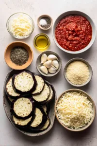 Crispy Baked Eggplant Parmesan Recipe (Authentic Italian Comfort Food Made Easy) 5 Flat lay of ingredients for baked eggplant parmesan including sliced eggplant, marinara sauce, mozzarella, parmesan, breadcrumbs, olive oil, and garlic