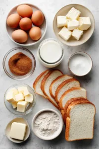Flat lay of ingredients for cinnamon sugar French toast recipe including sandwich bread, eggs, milk, cinnamon, sugar, vanilla extract, and butter on a light surface.