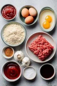 Ingredients for bbq meatloaf including ground beef, breadcrumbs, eggs, onion, garlic, barbecue sauce, ketchup, brown sugar, and spices arranged on a flat surface
