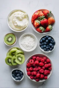 Ingredients for fruit pizza cookies including sugar cookie dough, cream cheese frosting, strawberries, blueberries, and kiwi arranged on a white surface