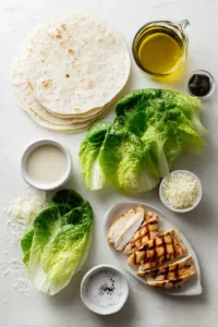 Easy Chicken Caesar Wrap Recipe (Quick & Healthy Dinner Idea) 5 Flat lay of ingredients for chicken caesar wrap including grilled chicken breast, romaine lettuce, flour tortillas, parmesan cheese, and Caesar dressing on a white background.