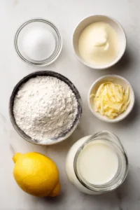 Ingredients for lemon mug cake including flour, sugar, milk, butter, lemon juice and zest on marble surface