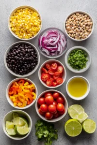 Flat lay of Texas caviar ingredients including black beans, black-eyed peas, corn, bell pepper, cherry tomatoes, red onion, lime, olive oil, and fresh cilantro.