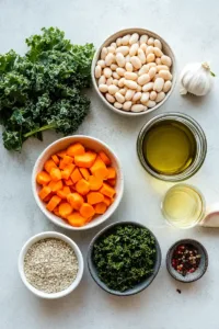 Flat lay of ingredients for Tuscan white bean soup including cannellini beans, chopped onion, garlic, carrots, celery, kale, olive oil, herbs, white wine, and vegetable broth.