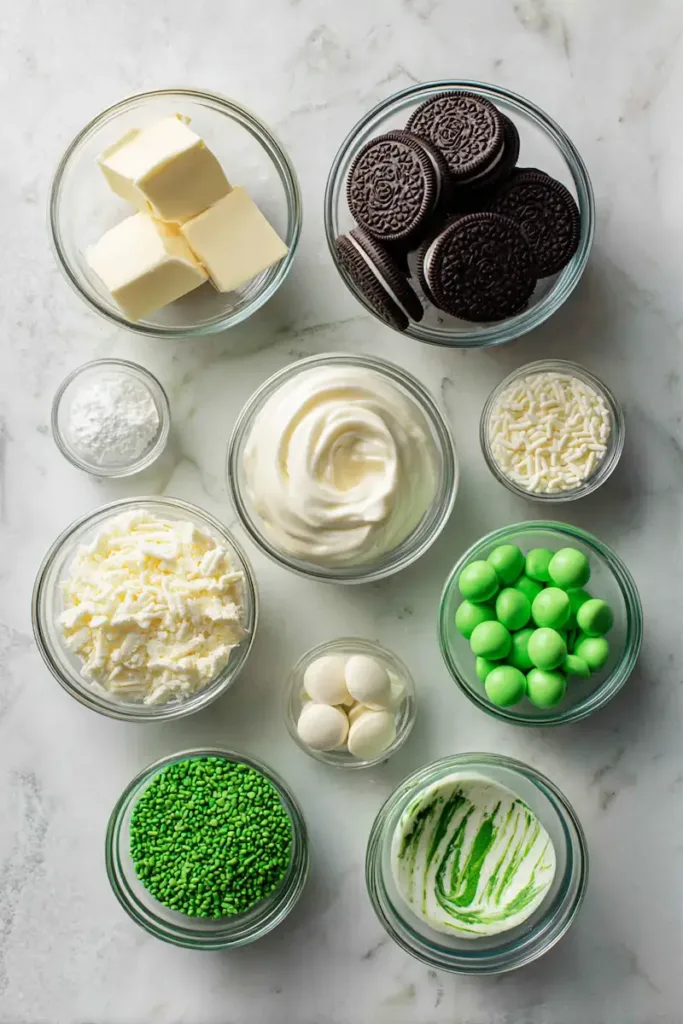Flat lay of ingredients for St. Patrick’s Day Oreo balls including chocolate sandwich cookies, cream cheese, white chocolate chips, green candy melts, and festive sprinkles on a light surface.