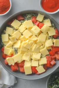 Flat lay of ingredients for spicy Velveeta spinach dip including Velveeta cheese, fresh spinach, Rotel tomatoes, cream cheese, jalapeño, garlic, and spices on a white surface