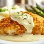 Crispy parmesan crusted chicken with creamy garlic sauce served on a white plate with mashed potatoes and roasted asparagus, easy family dinner recipe.