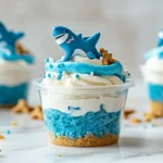 No-bake Shark Week cheesecake cups in clear dessert cups with blue topping, gummy sharks, and cookie crumb sand.
